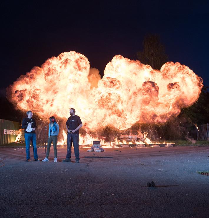 Three people standing in front of a large explosion at night time