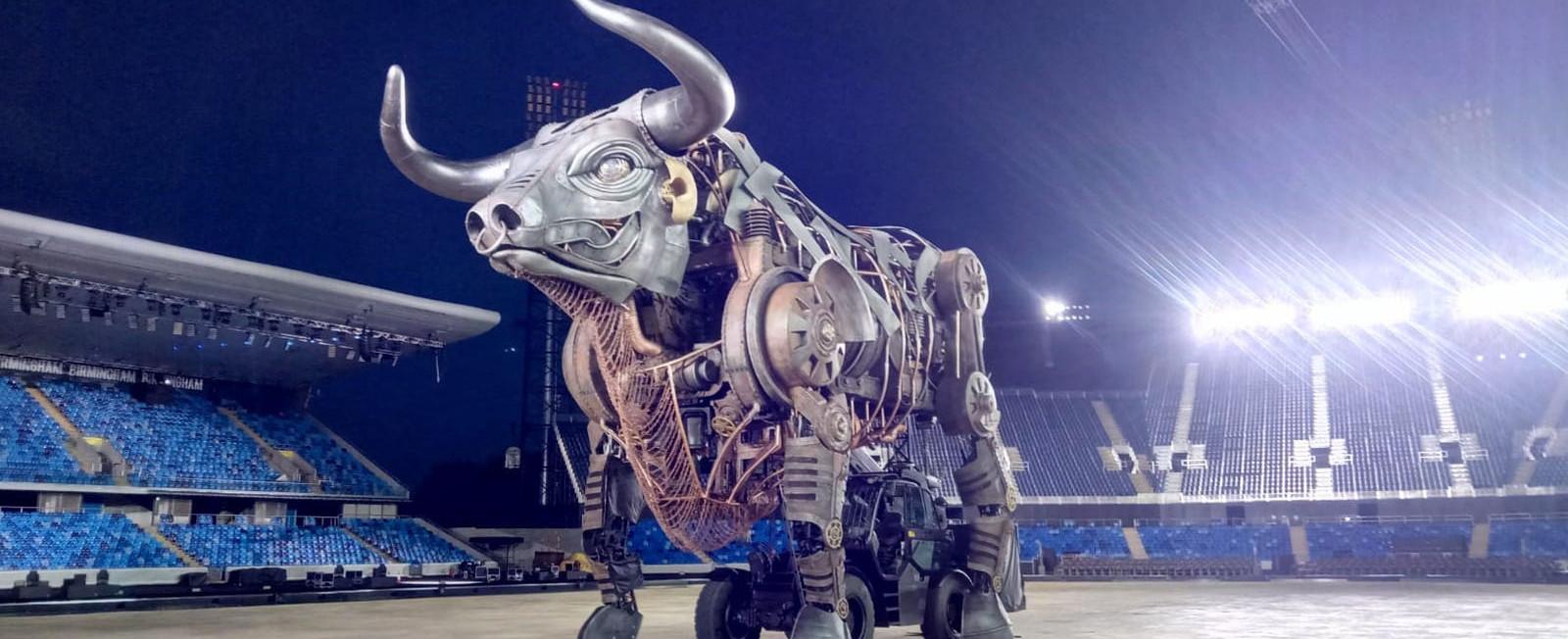 A large metal steampunk style mechanical bull in the middle of an empty stadium at light, lit up by the stadium lights