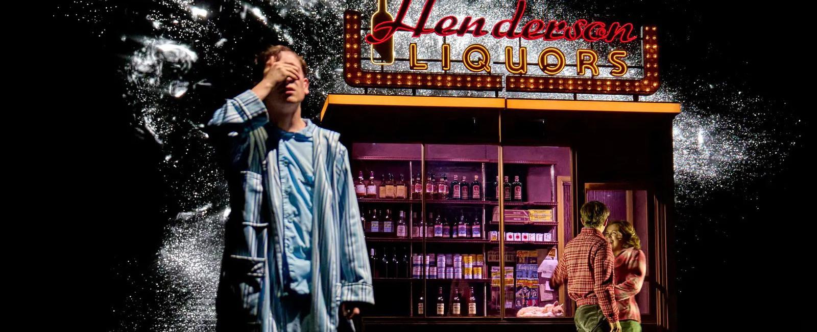 A production still from 'Stranger Things The First Shadow - Henry Creel in the foreground with his hand covering his face, and two cast members chatting in front of a liquor store in the background